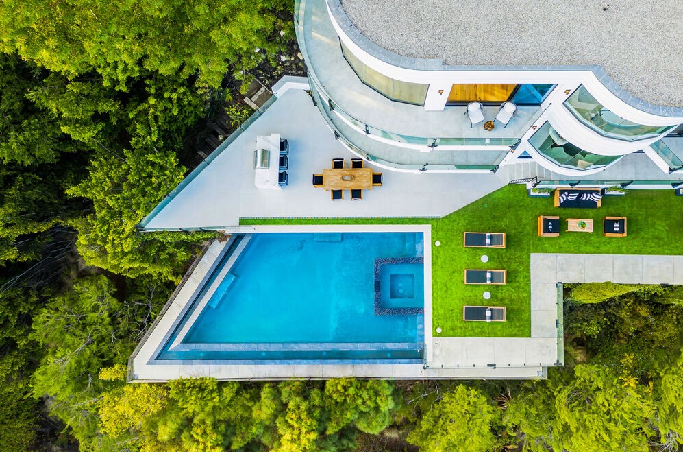 Aerial view of a curved modern hillside home with an infinity pool and terraces set into the trees